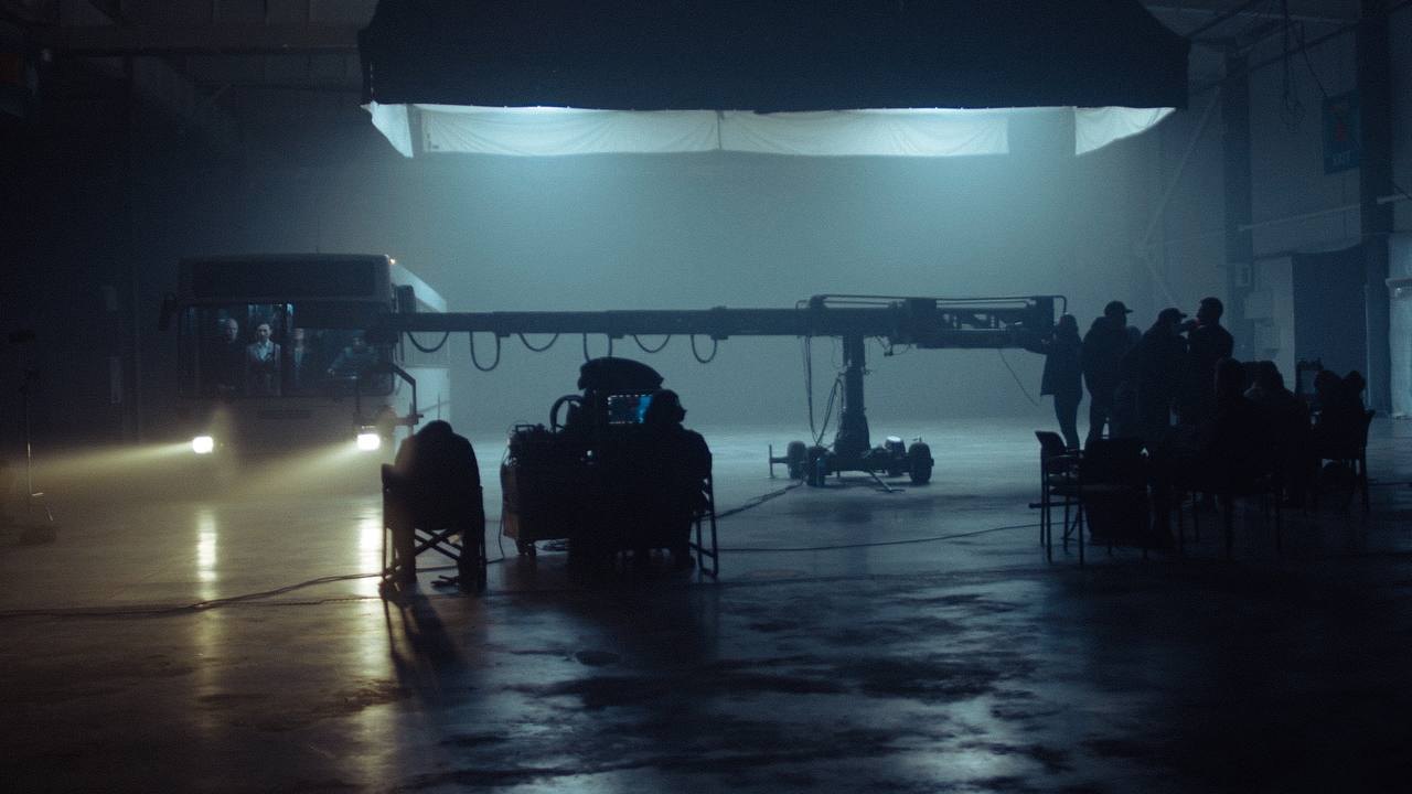 Wide shot of the set: bus with headlights on, camera crane and crew chairs in haze