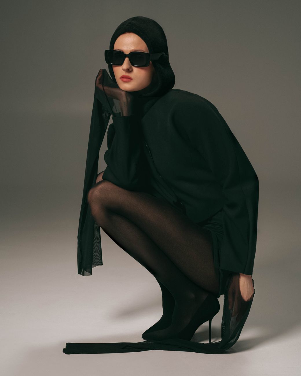 Model in black with sunglasses crouching on a grey studio floor
