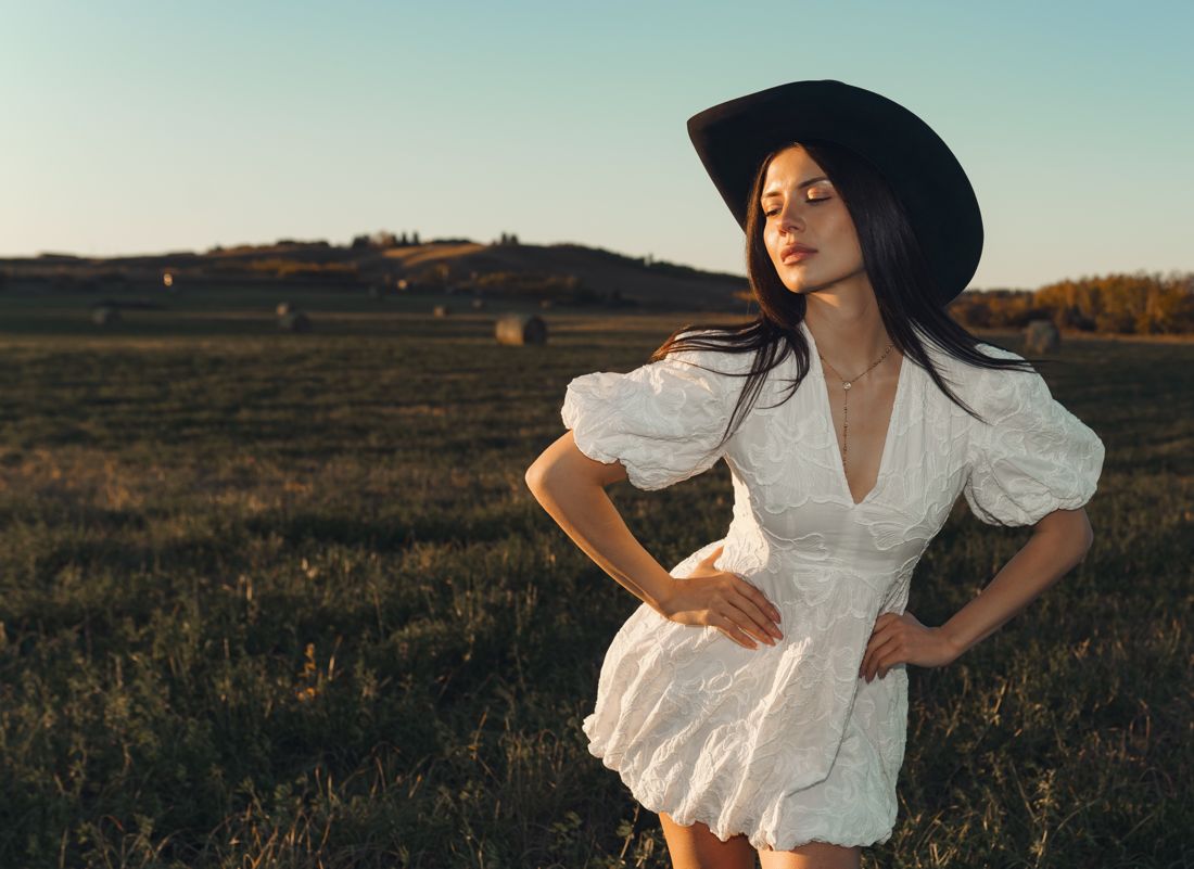 Model in a white dress and black cowboy hat in an Alberta field, hands on hips